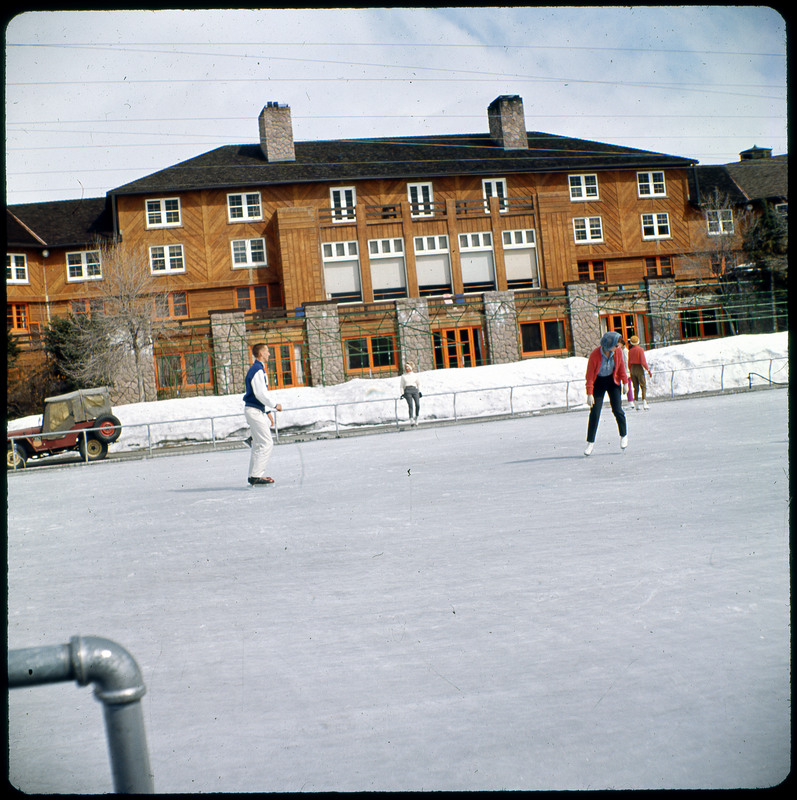 A photographic slide of an ice skating rink. There are people skating and a lodge in the background.