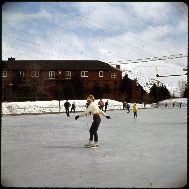 A photographic slide of an ice skating rink. There is a woman skating by in the foreground, and many people skating in the background. There is also a lodge in the background.