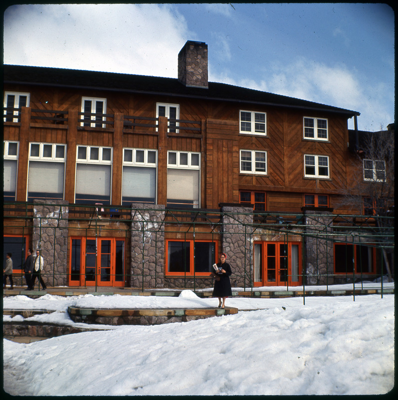 A photographic slide of Evelyn Crabtree standing in front of a lodge. There is snow on the ground and blue sky.