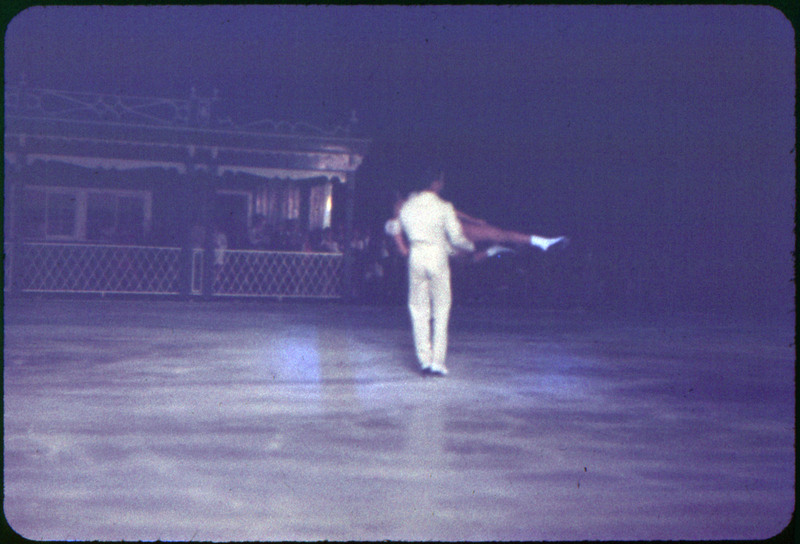 A photographic slide of a man in a white suit holding a woman horizontally. They seem to be ice skating for a crowd.