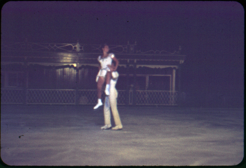 A photographic slide of a man holding a woman up in the air during an ice skating dance. There is a crowd in the background.