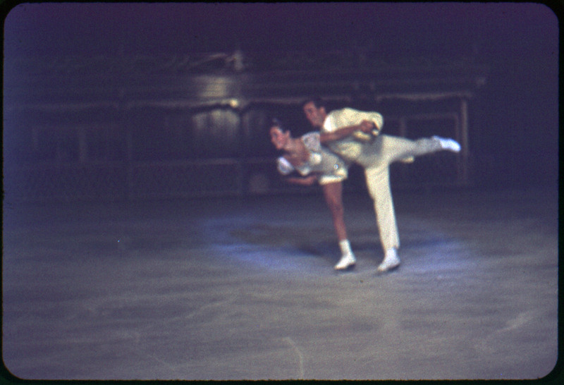 A photographic slide of a couple ice skating hand in hand and both on one leg. They are in a white costume and there are people in the background.
