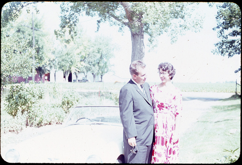 A photographic slide of a couple standing in front of a car in a park. The woman is wearing a purple dress and they are looking at one another.