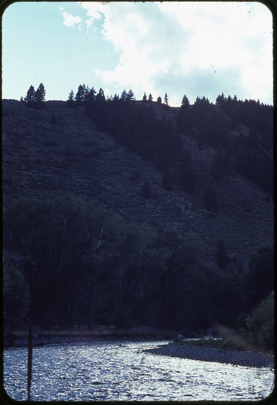 A photographic slide of a river valley. There are trees at the base of a hillside and what seems like a person fishing on riverside.