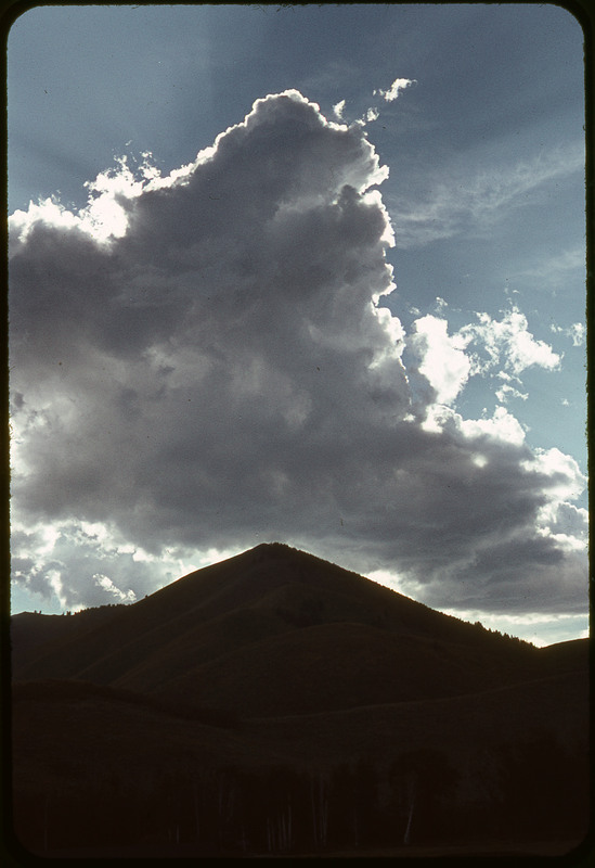 A photographic slide of a dark mountain with giant blue skies and clouds above them.