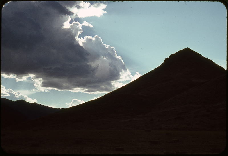 A photographic slide of a mountain range that is dark. There is a blue sky and giant clouds.