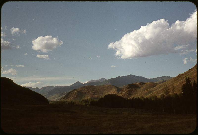 A photographic slide of a giant mountain range. There is a blue sky and clouds. There is no snow.