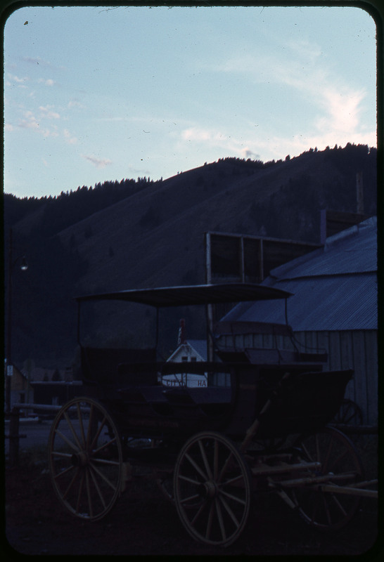 A photographic slide of a carriage parked next to a building. There are mountains in the background.