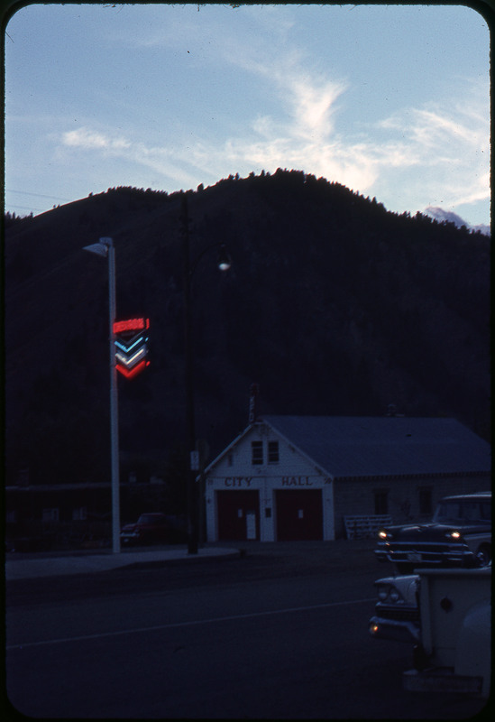 A photographic slide of a city hall building in front of a mountain. There are cars driving on the road nearby.