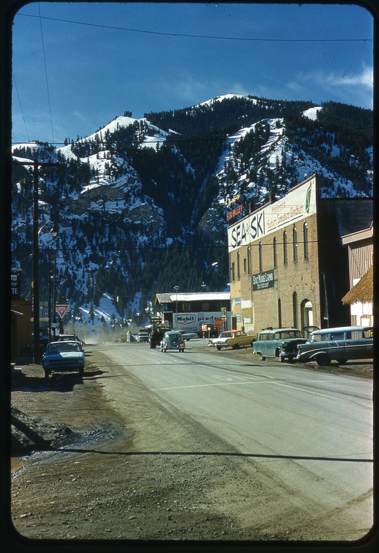 A photographic slide of a snowy mountain in the background. There is a road with cars on it and a small town of buildings.
