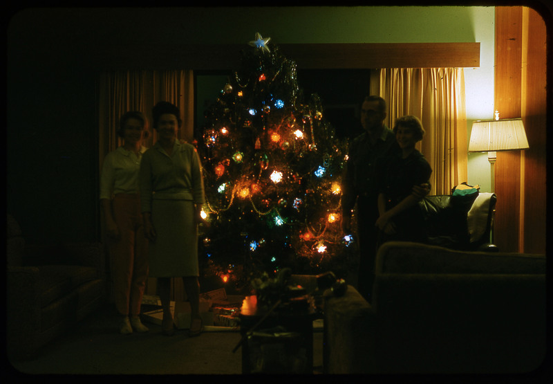 A photographic slide of a four person group standing next to a Christmas tree. It is lit up and there are presents underneath.