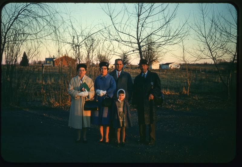 A photographic slide of a family standing outside in front of a agricultural field. They are dressed in nice clothes. There are houses and barns in the background.
