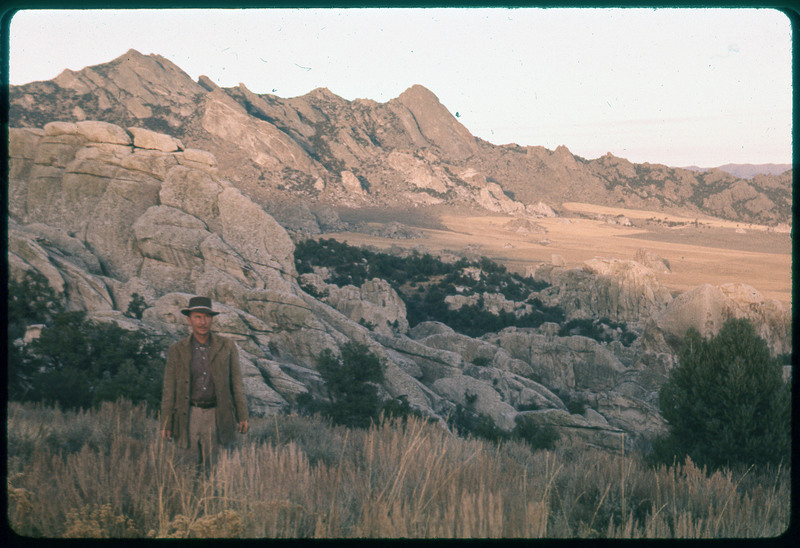 A photographic slide of Donald Crabtree posing in front of a rocky valley. The mountains are rocks and there are some fields in the background.