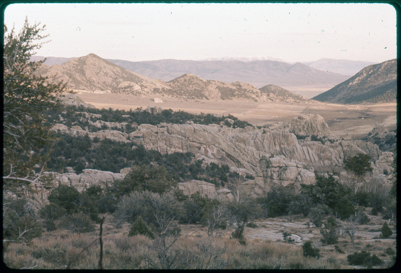 A photographic slide of a rocky valley with many mountains in the background. There are also many trees and bushes.