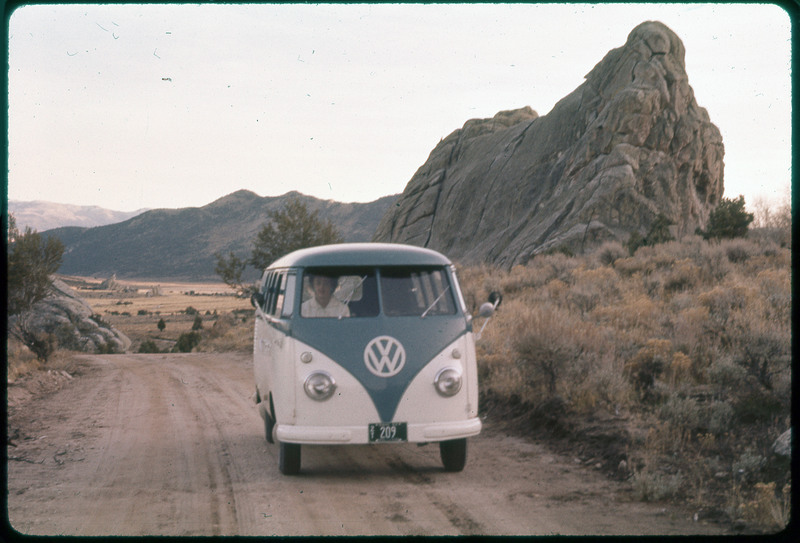 A photographic slide of Evelyn Crabtree driving a VW van on a dirt road. There are large rock formations in the background and mountains.
