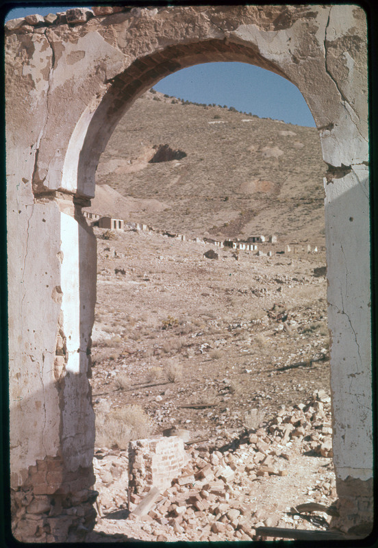 A photographic slide of an old archway from a structure. There are other old structures in the background and a mountain.