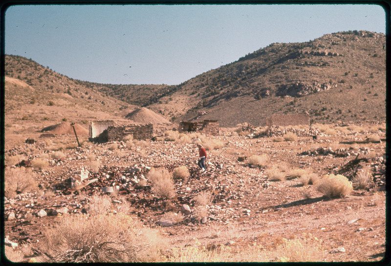 A photographic slide of a mountain with an old structure at the base. the mountain is small, rocky, and has bushes.