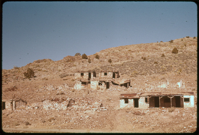 A photographic slide of brick structures built at the base of a rocky hill. There are some trees.