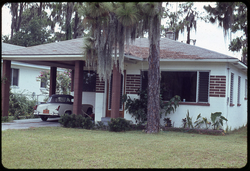A photographic slide of a nice white and red brick house with a car in a car park. There is a lawn in front with large trees.