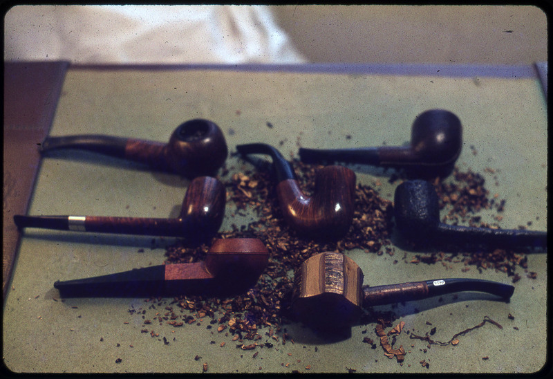 A photographic slide of seven pipes carved and sitting on a surface. There are piles of wood chips underneath the pipes.
