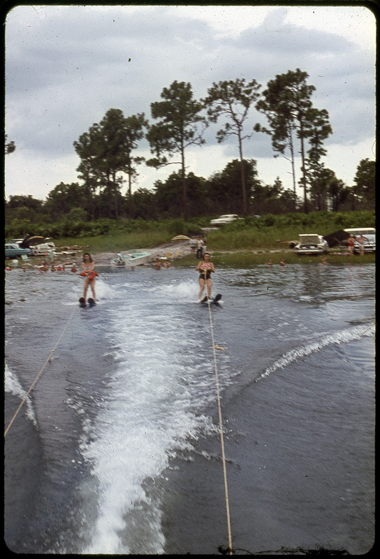 A photographic slide of two people water skiing. There is a beach in the background with cars parked and people on the beach.
