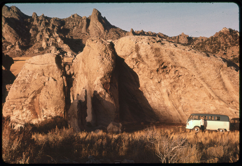 A photographic slide of a rock formation. Evelyn Crabtree and the VW van are parked in front of the rock. There are rocky mountains in the background.