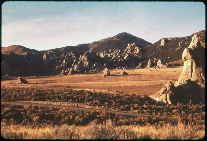 A photographic slide of rocky landscape with mountains and a road. There are multiple rock structures in a field.