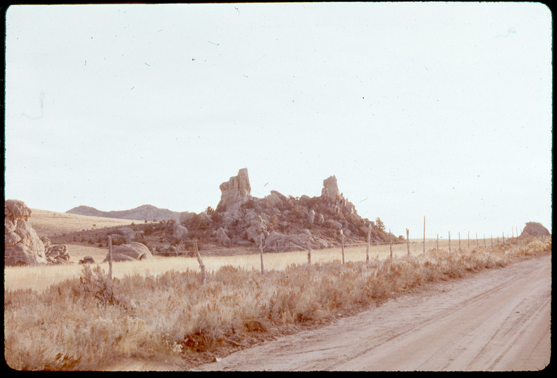 A photographic slide of a rocky formation behind a fence and along a road. There are mountains in the background.