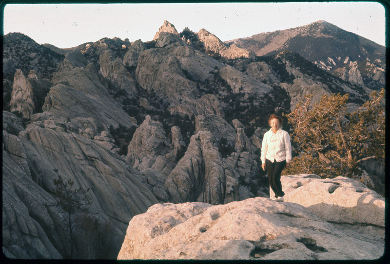 A photographic slide of a rocky mountain with Evelyn Crabtree standing in front. The rocks are in unique formations and there are mountains in the background.
