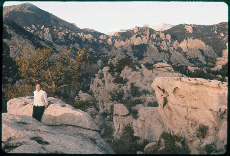 A photographic slide of Evelyn Crabtree standing in front of rocky mountain range. There are unique rock formation and a mountain range in the background.