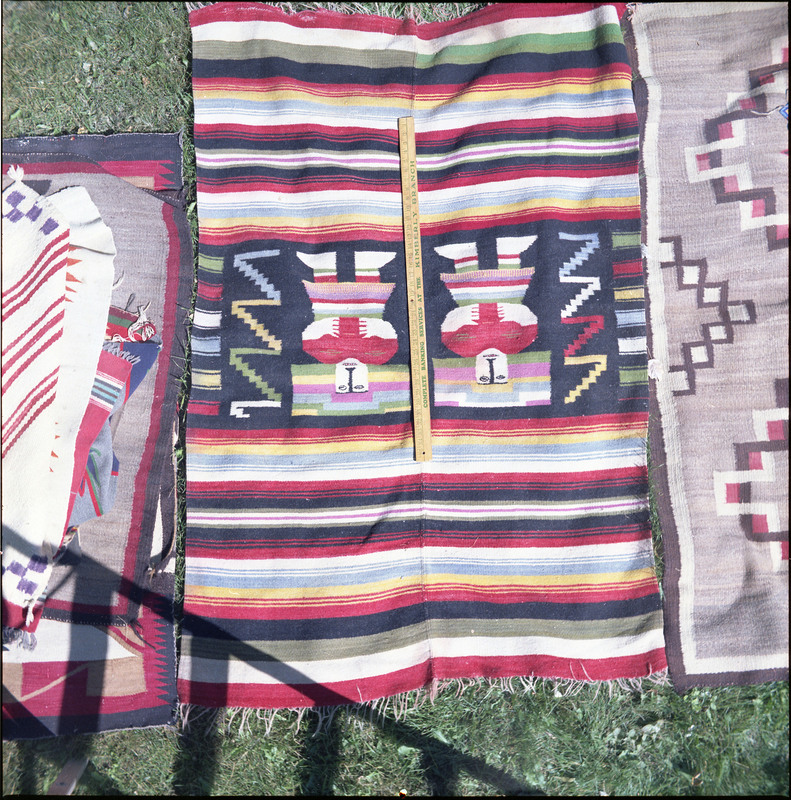 An image of many woven blankets created by unknown creators with a yard stick on one. Two blankets are laid out and there is a pile of more woven blankets on the left of the image.
