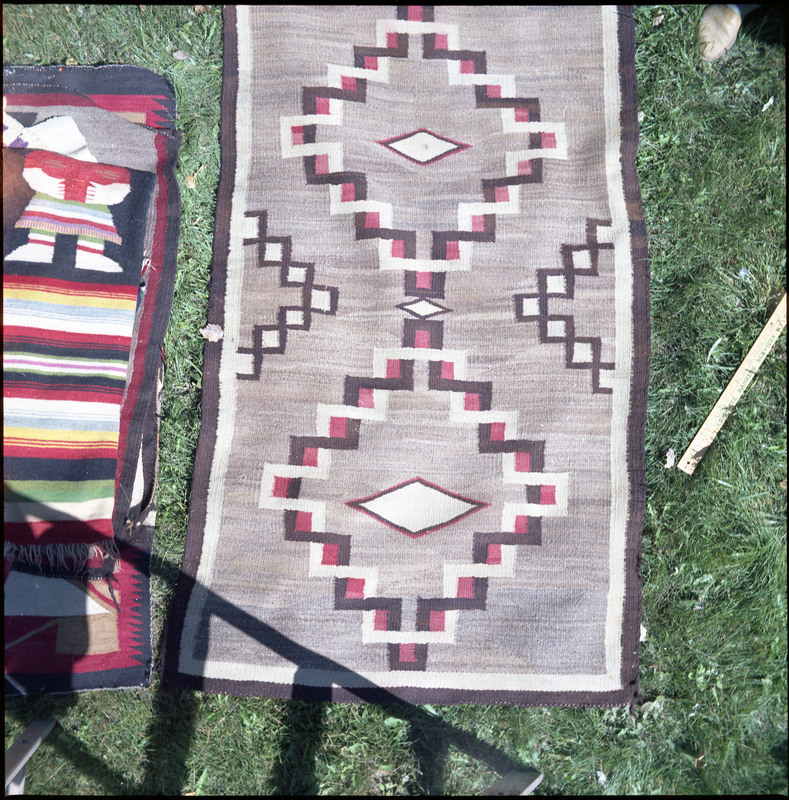 An image of a woven blankets created by an unknown creator. One blanket is laid out and there is a pile of more woven blankets on the left of the image.