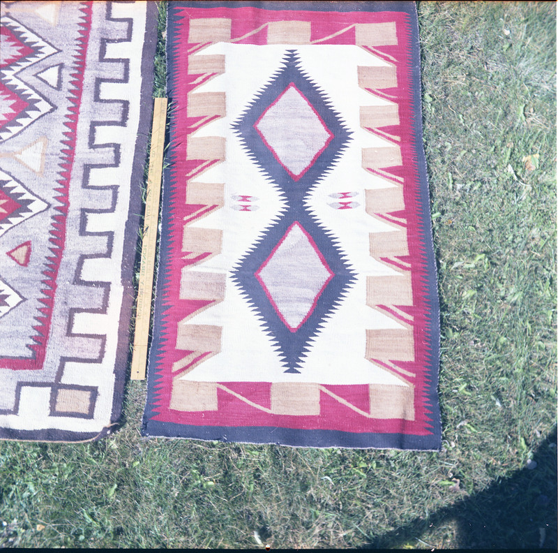 An image of many woven blankets created by unknown creators with a yard stick to indicate size. Two blankets are laid out side by side.