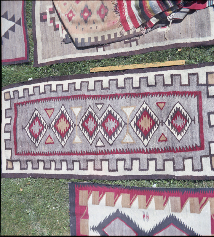 An image of many woven blankets created by unknown creators with a yard stick to indicate size. Two blankets are laid out side by side and a pile of woven items is present in the top portion of the image.