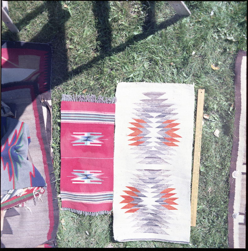 An image of many woven blankets created by unknown creators with a yard stick on one. Two blankets are laid out and there is a pile of more woven blankets on the left of the image.