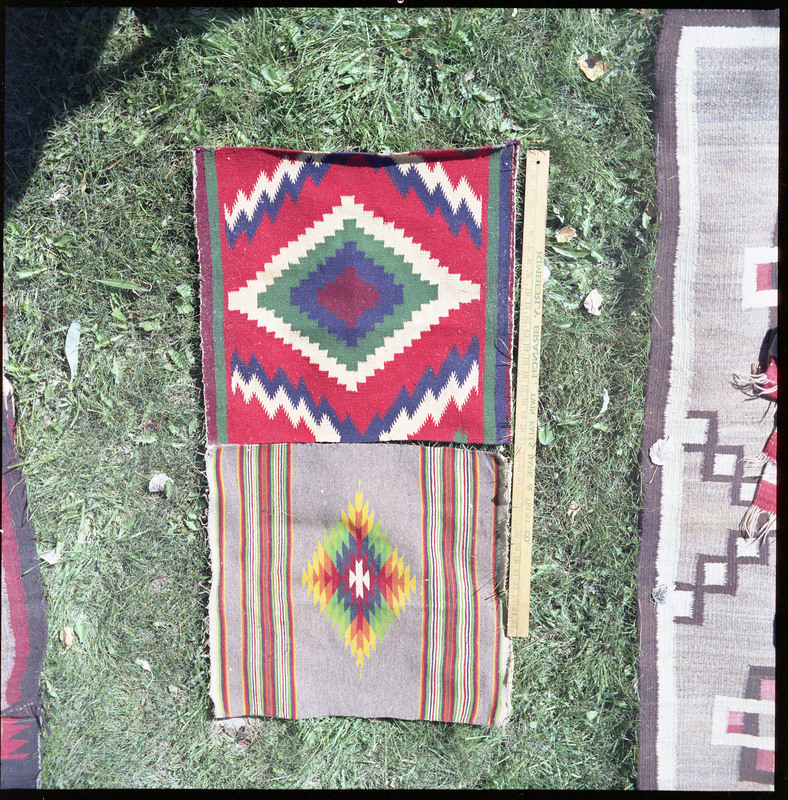 An image of many woven blankets created by unknown creators. Two blankets are laid out side by side with a yard stick indicating size.