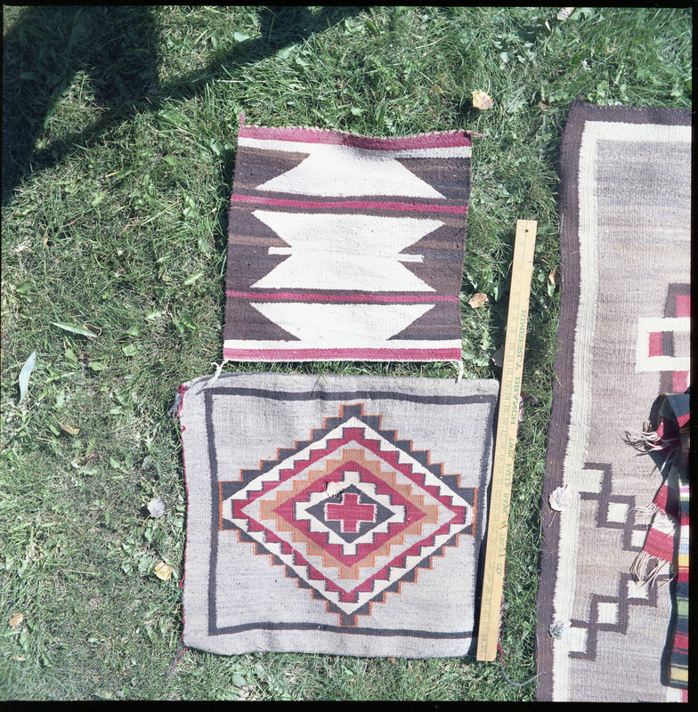 An image of many woven blankets created by unknown creators. Two blankets are laid out side by side with a yard stick indicating size.The blankets are small, and patterned with vibrant colors.