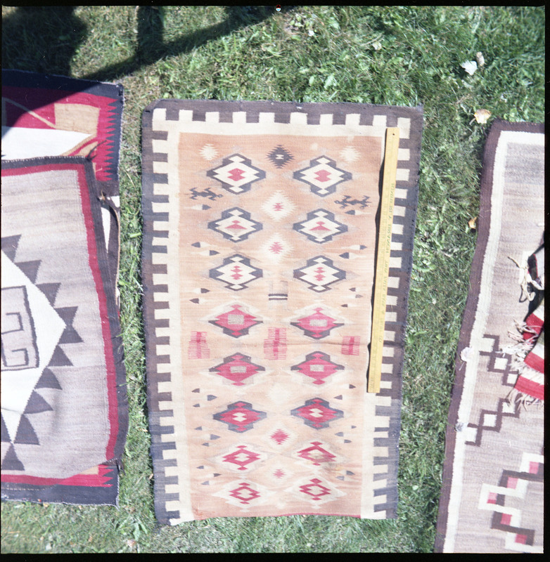 An image of many woven blankets created by unknown creators with a yard stick on one. One blanket is laid out as the focus of the image and there are more woven items to the left and right of the image.