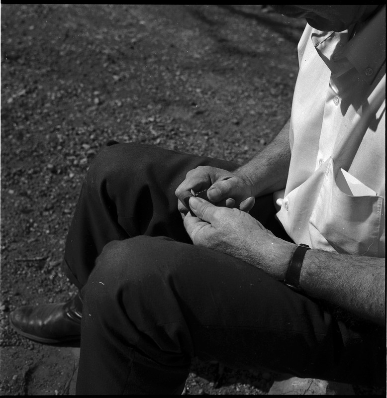 An image of Donald E. Crabtree flintknapping a small arrowhead. Crabtree is seated on a log, working on the stone object in his hands.