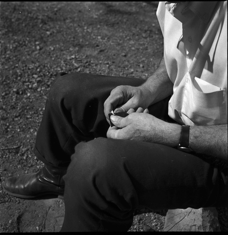 An image of Donald E. Crabtree flintknapping a small arrowhead. Crabtree is seated on a log, working on the stone object in his hands.