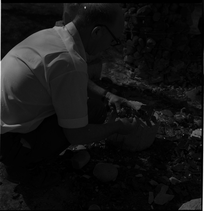 An image of Donald E. Crabtree looking at stone objects that are resting on a large rock. This image is dark and difficult to descirbe.