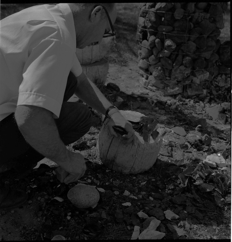 An image of Donald E. Crabtree looking at stone objects that are resting on a large rock. It appears as though he is preparing to use a hammerstone in his right hand to strike the stone he is holding in his left hand, resting against a log stump.
