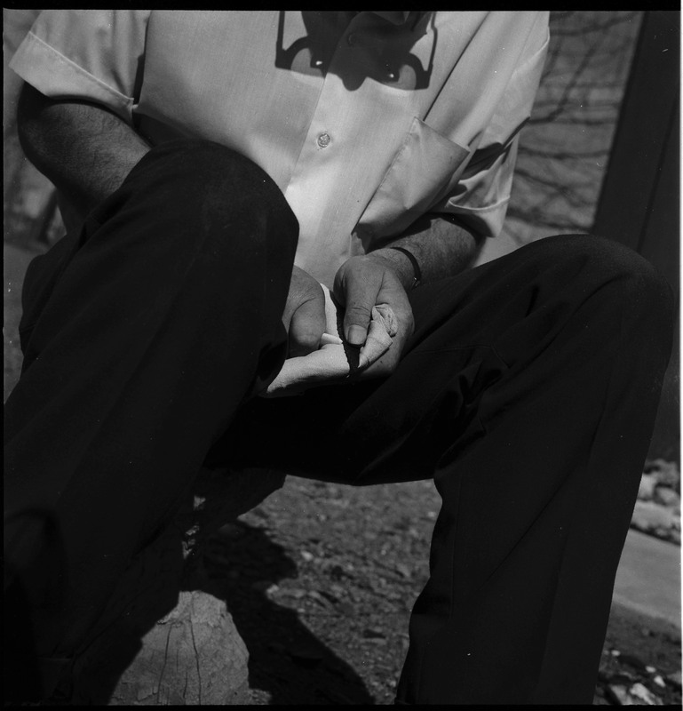 An image of Donald E. Crabtree flintknapping a small arrowhead. Crabtree is seated on a log, working on the stone object in his hands. In this image he is using a small item, possibly bone, to pressure flake the object he is holding in a cloth.