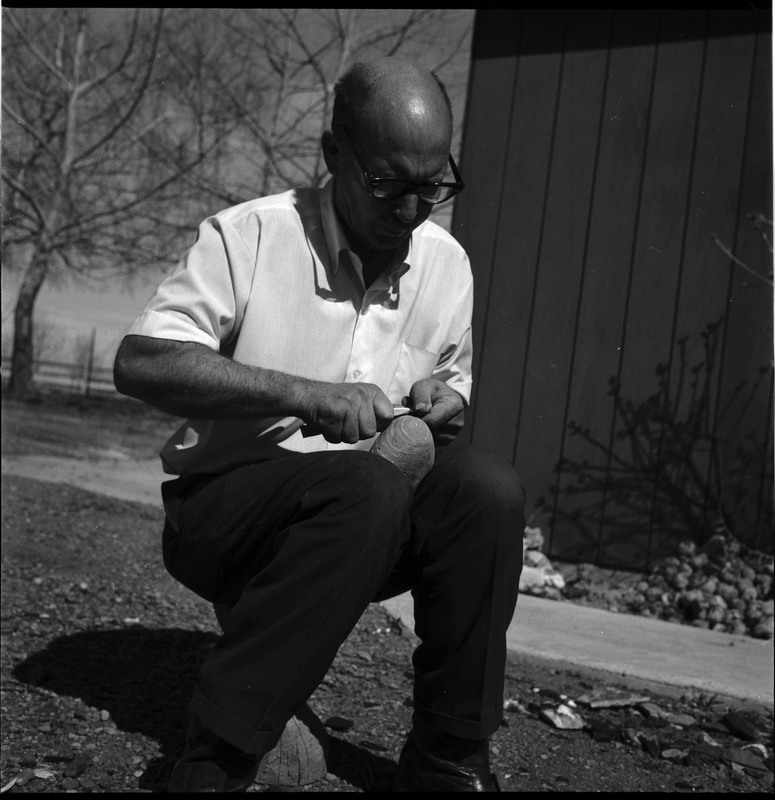 An image of Donald E. Crabtree flintknapping a small arrowhead. Crabtree is seated on a log, working on the stone object in his hands. In this image he is using a small item, possibly bone, to pressure flake the object he is holding.