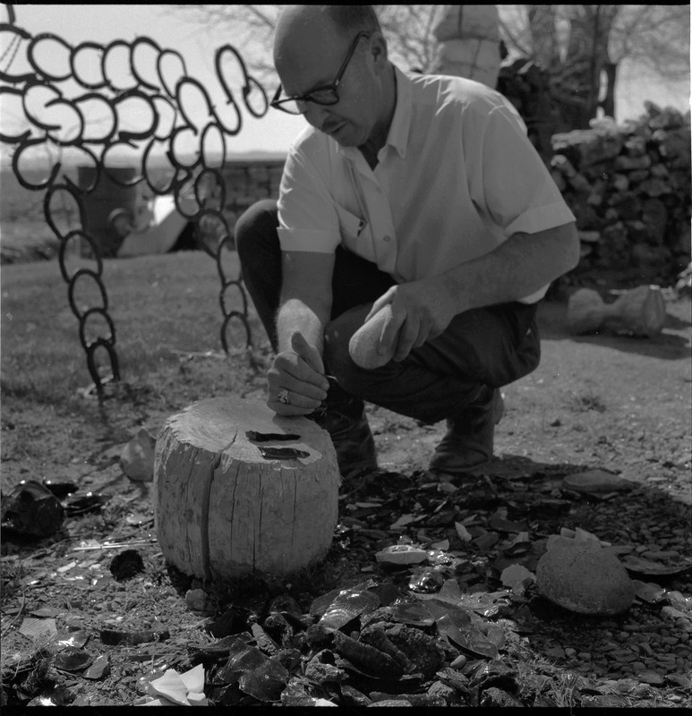 An image of Donald E. Crabtree getting ready to strike a core that appears to be obsidian. There are obsidian flakes resting on the log stump. Crabtree is outside and there is a horseshoe sculpture in the background.