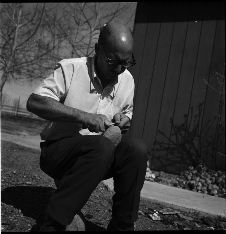 An image of Donald E. Crabtree flintknapping a piece of obsidian. Crabtree is seated on a log, working on the stone object in his hands. In this image he is using a small item, possibly bone, to pressure flake the object he is holding.