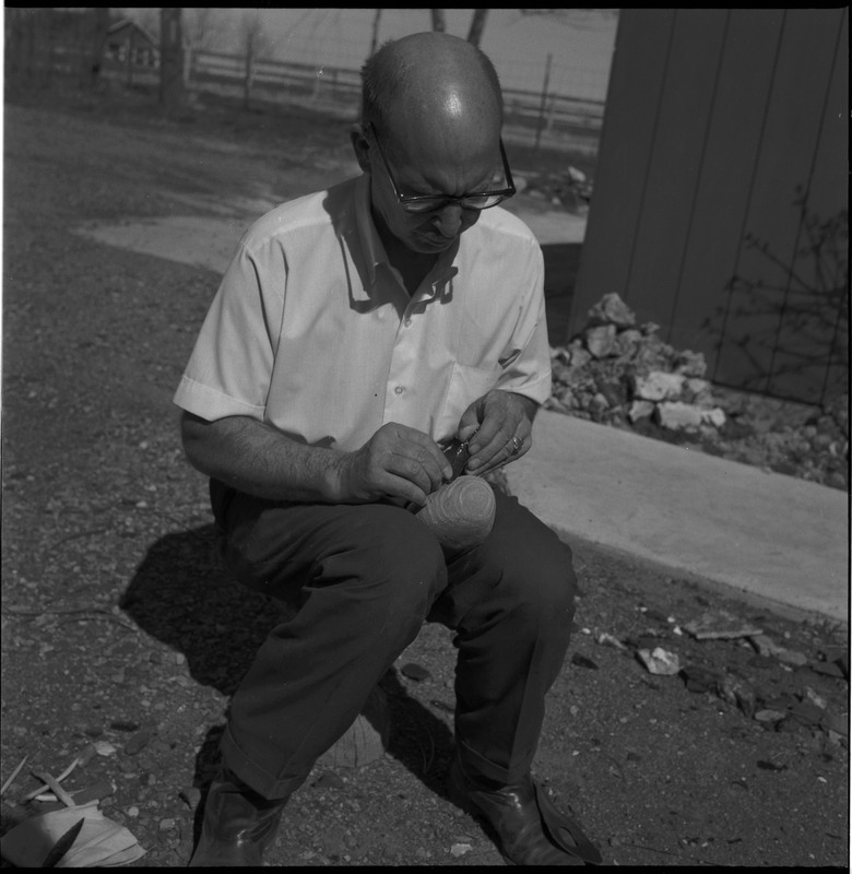 An image of Donald E. Crabtree flintknapping a piece of obsidian. Crabtree is seated on a log, working on the stone object in his hands. In this image he is using a small item, possibly bone, to pressure flake the object he is holding.