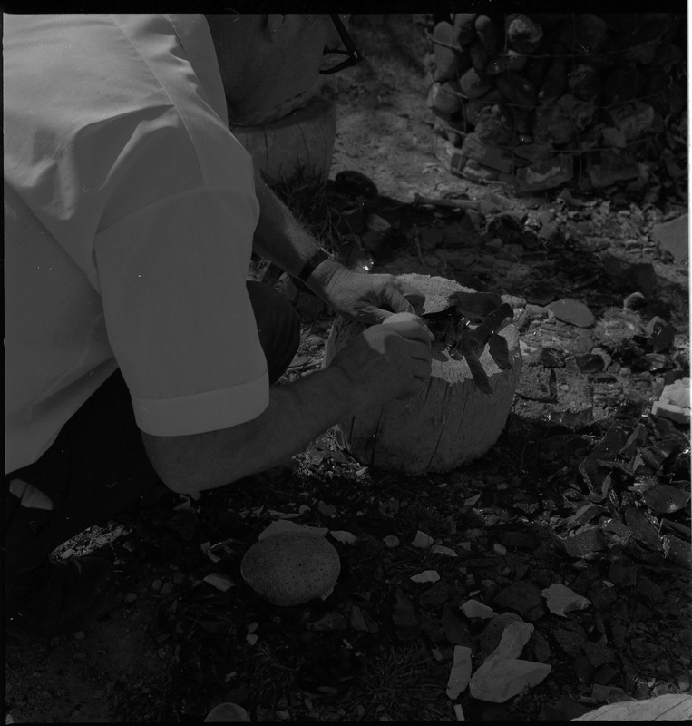 An image of Donald E. Crabtree flintknapping on a log stump. He is working on blades of what looks like obsidian. He is in a working area, there are pile of debitage surrounding the log stump.