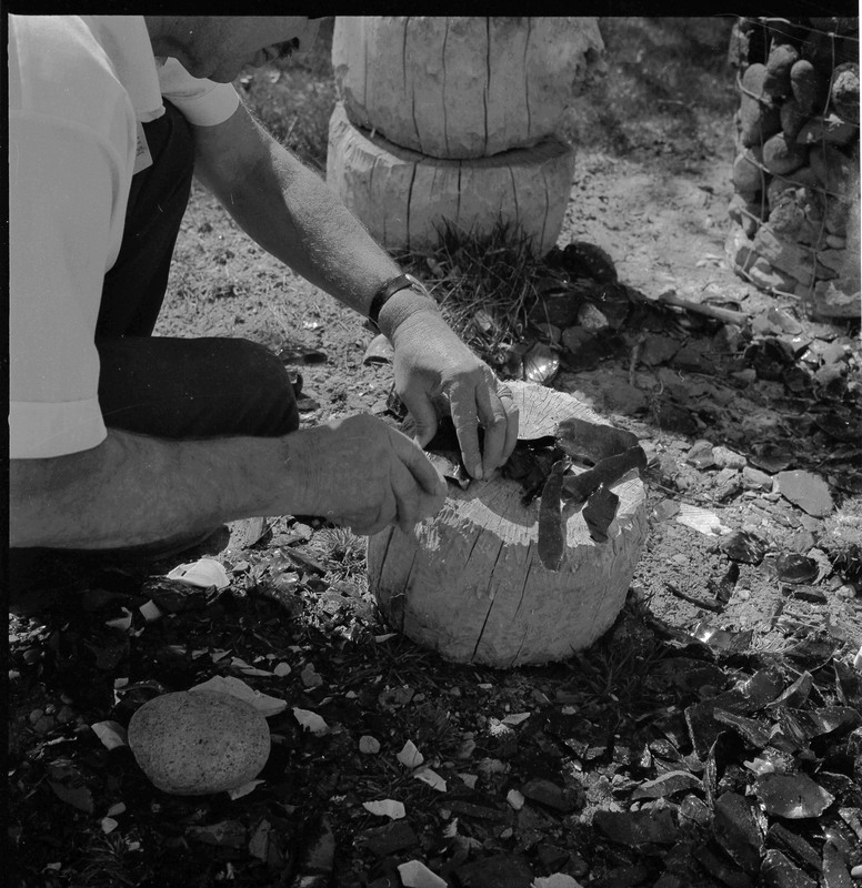 An image of Donald E. Crabtree flintknapping on a log stump. He is working on blades of what looks like obsidian. He is in a working area, there are pile of debitage surrounding the log stump.