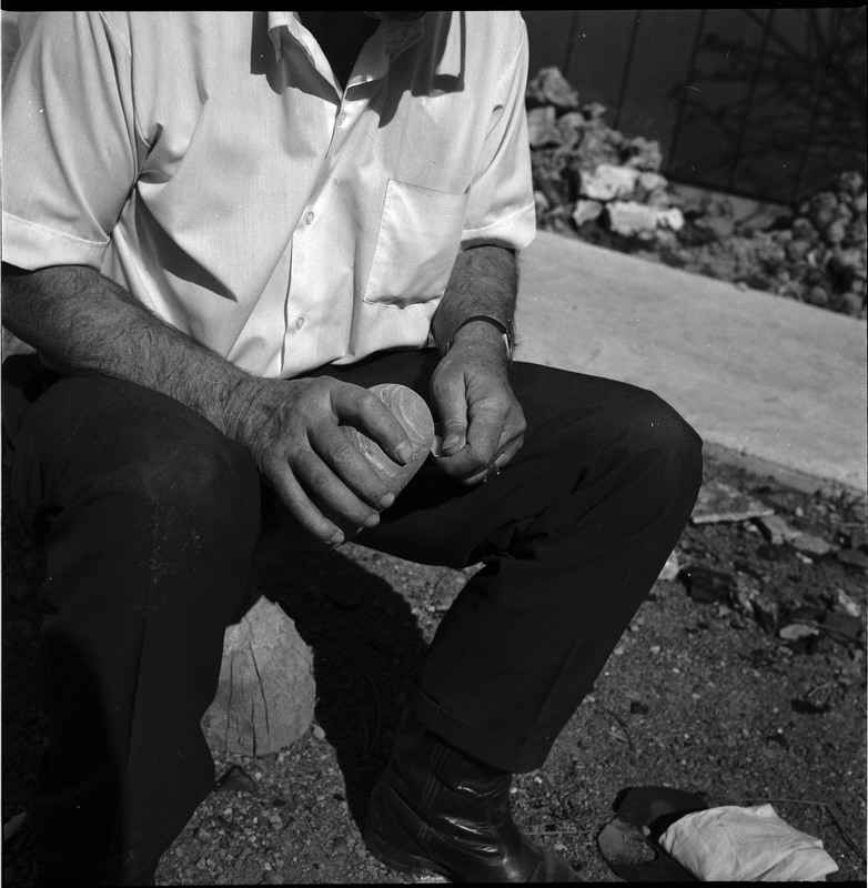 An image of Donald E. Crabtree flintknapping on a log stump. Crabtree is using a large hammerstone to ground the edges of the item in his hands.